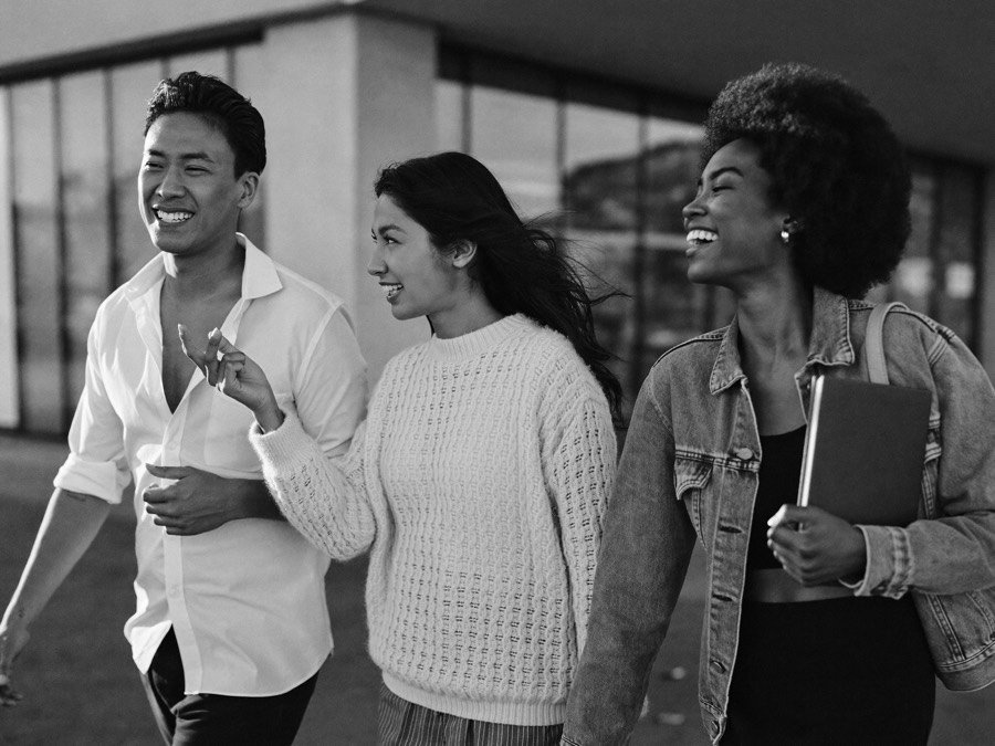 Three friends walking and talking outdoors, one explaining an idea with her hands while the others listen and laugh.