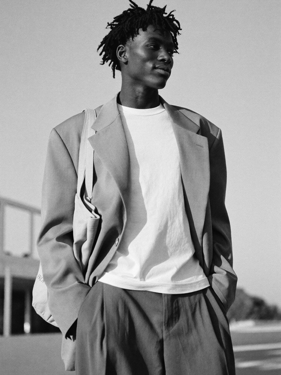 A young man in an oversized blazer and white t-shirt, a tote on his shoulder, looking toward the horizon in low sun.