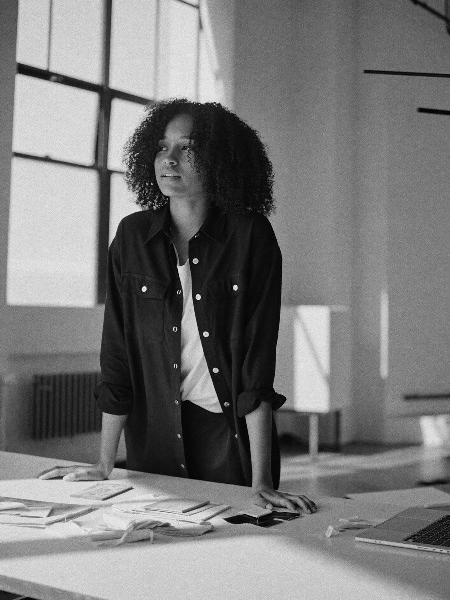 A young designer with curly hair at a studio worktable, one hand on pinned sketches, looking toward the window light.
