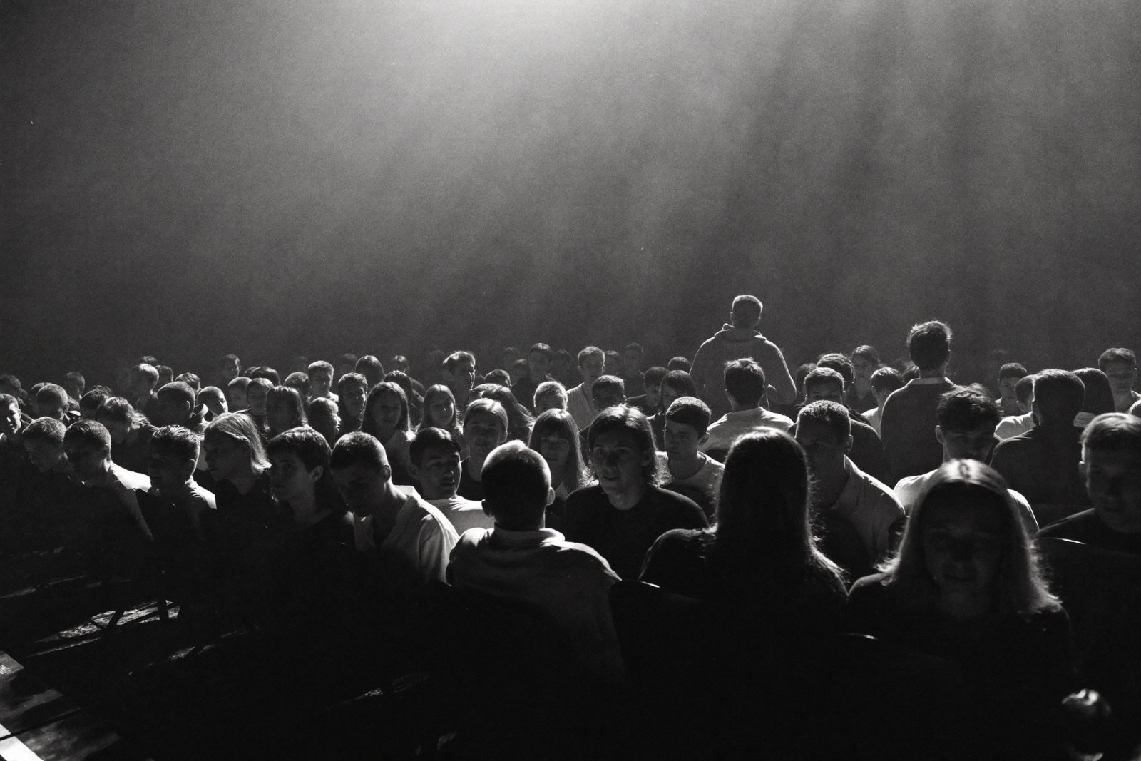 A packed auditorium seen from the edge of the stage, faces lit by a beam of light through haze.