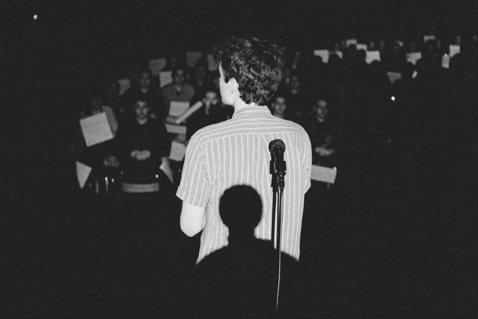 A founder seen from behind at the centre of a stage, facing a full auditorium, a microphone in front of him.
