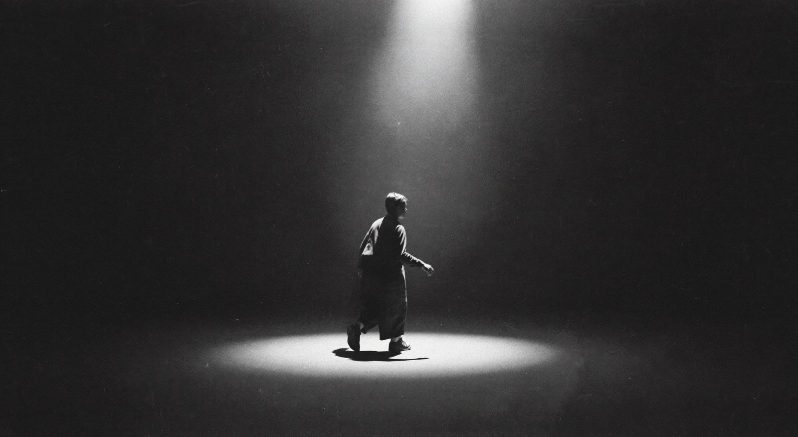 A lone figure walks out of the darkness into a single spotlight at the centre of a vast empty stage.